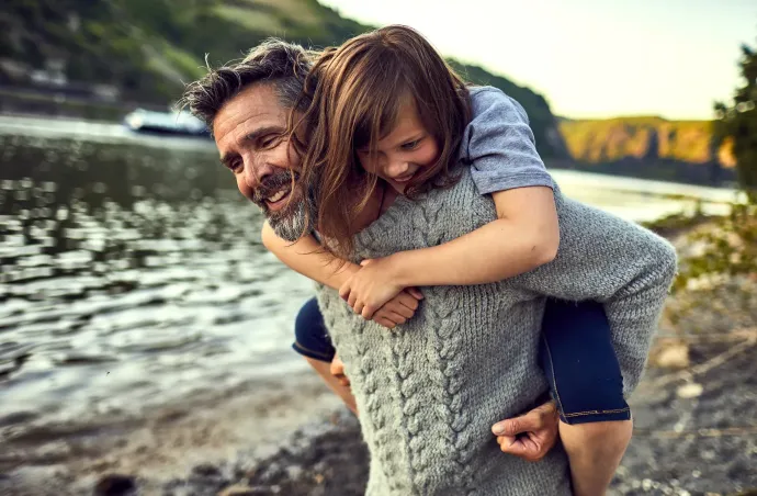 a father with travel insurance carries a child on their back while walking along the shore of a lake