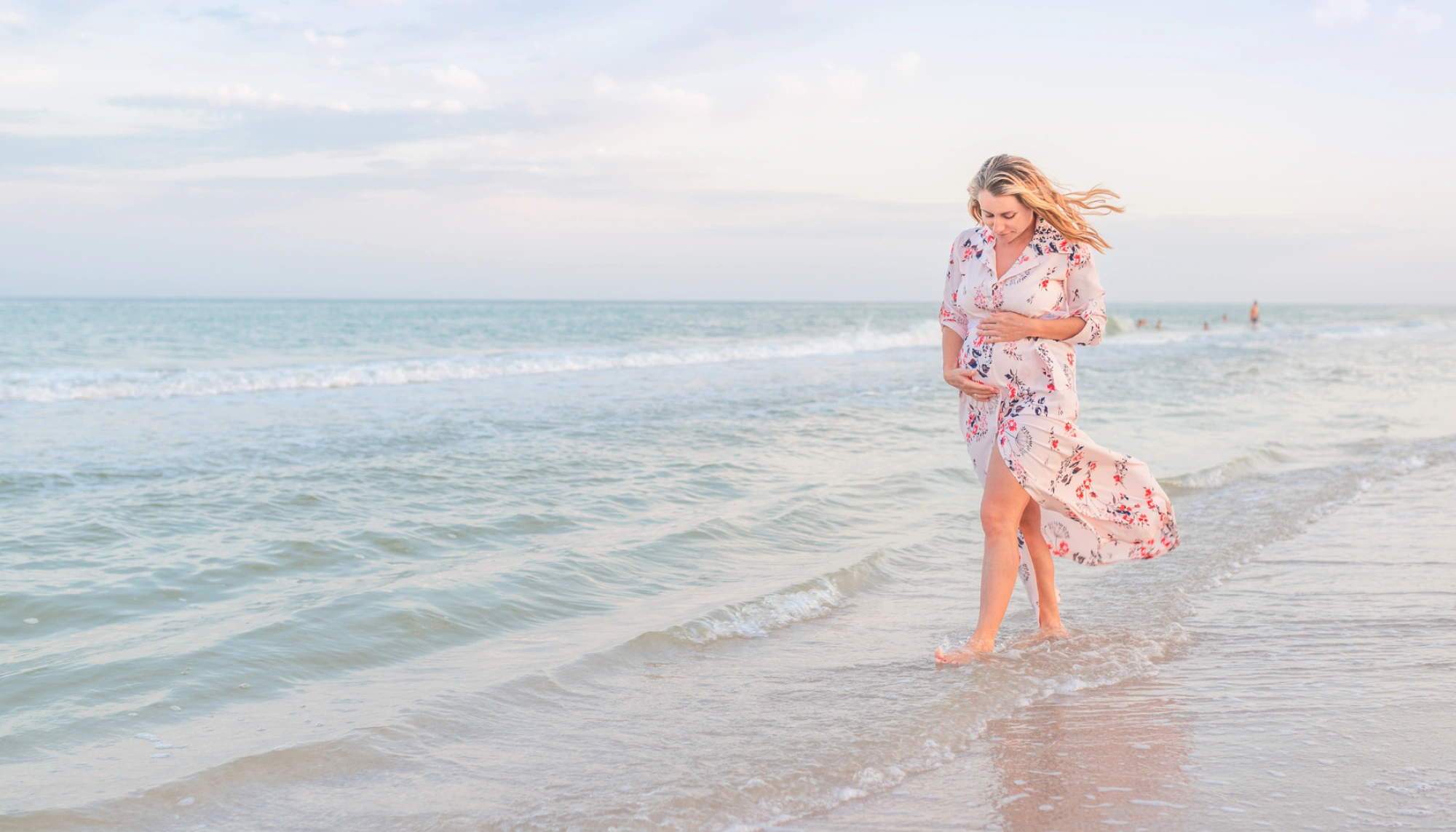a pregnant woman in a floral dress walks on the beach on vacation after deciding to travel during pregnancy