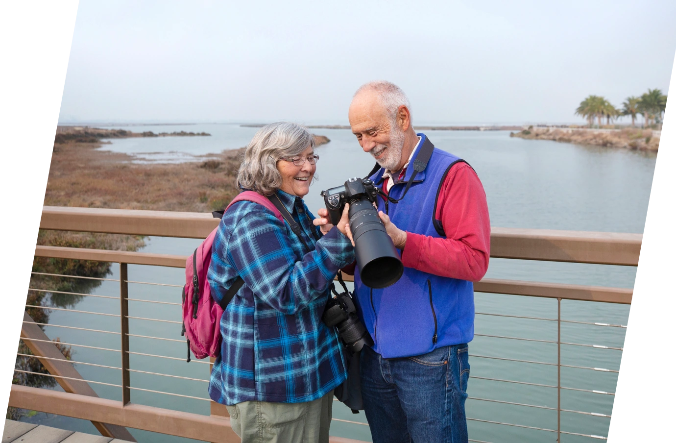 an older couple with travel insurance for pre-existing conditions reviews a camera's photos on a wooden bridge over a marsh