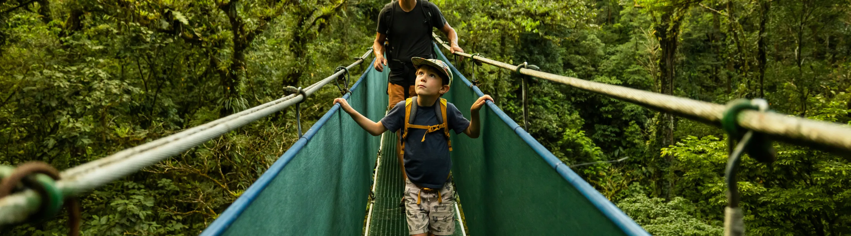 a father and son cross a suspension bridge in a tropical rainforest, insured with travel insurance for Costa Rica 