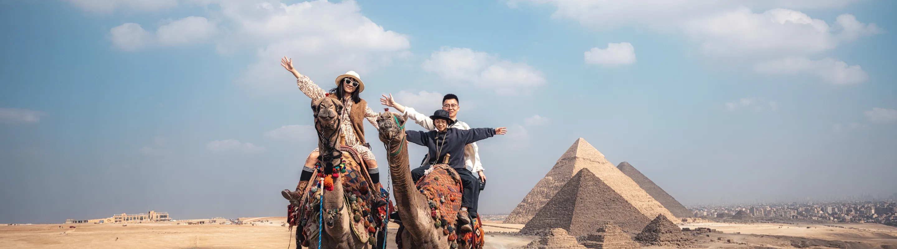 a family of three raises their arms as they ride camels at the Pyramids of Giza, insured with travel insurance for Egypt