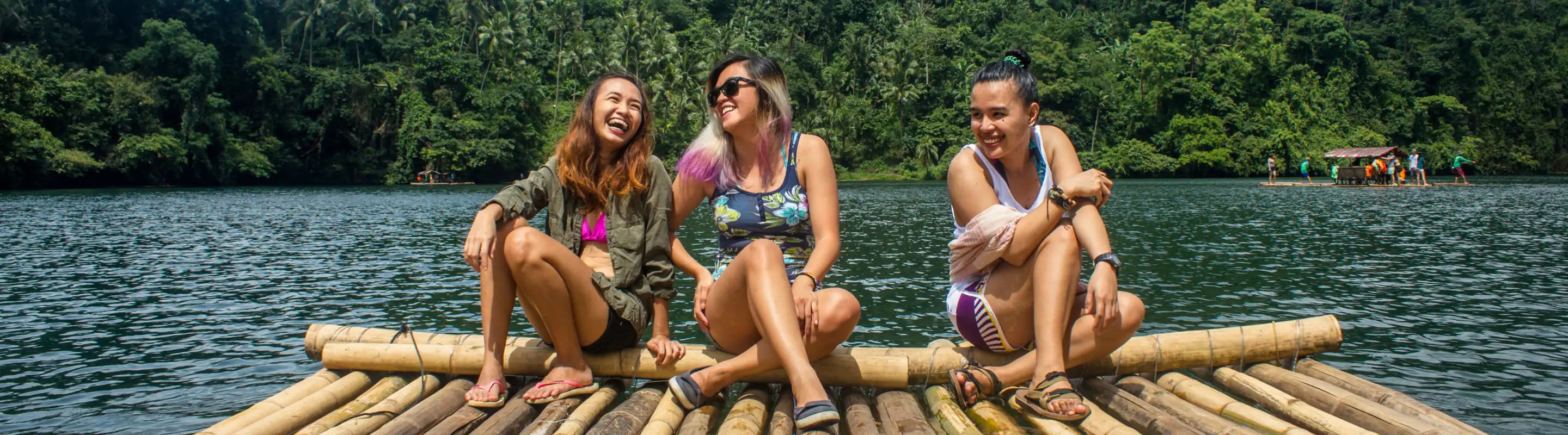 three young women sitting on a bamboo raft in the water, insured with travel insurance for the Philippines