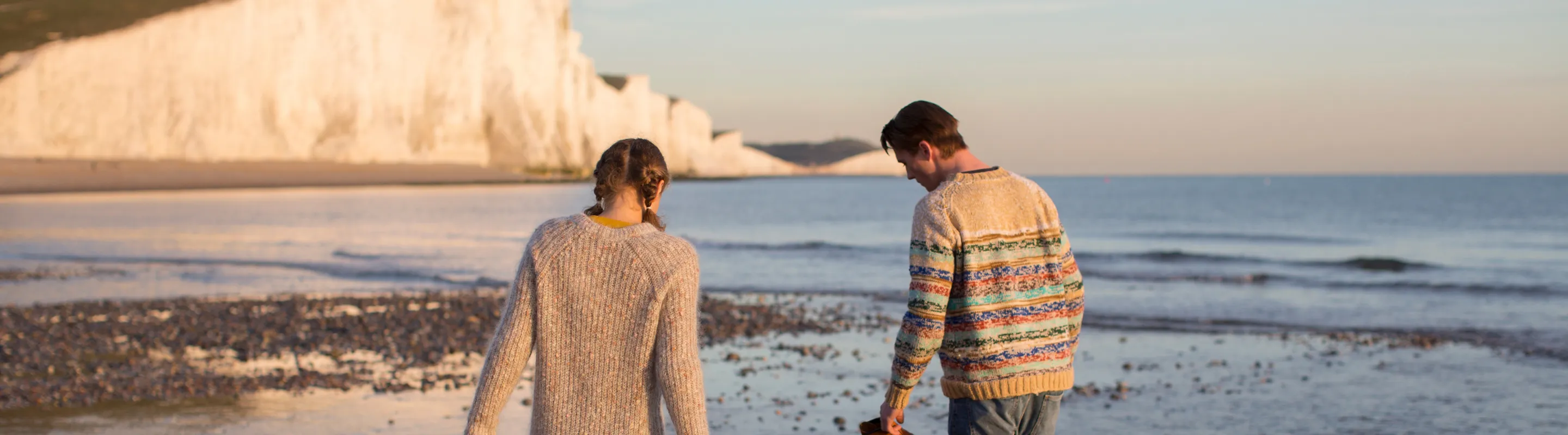 two travelers with travel insurance for the UK walk barefoot along a pebble beach beneath the Seven Sisters white cliffs