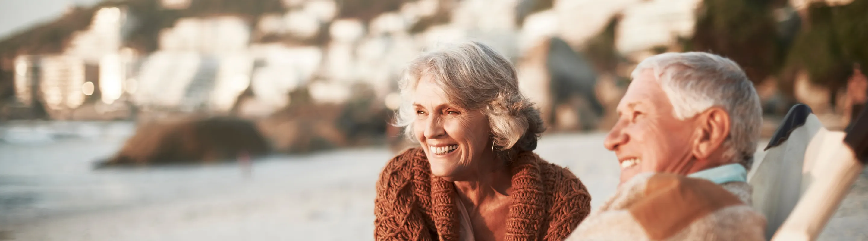 two older adults smile and sit in beach chairs along a shoreline on vacation after comparing travel insurance plans