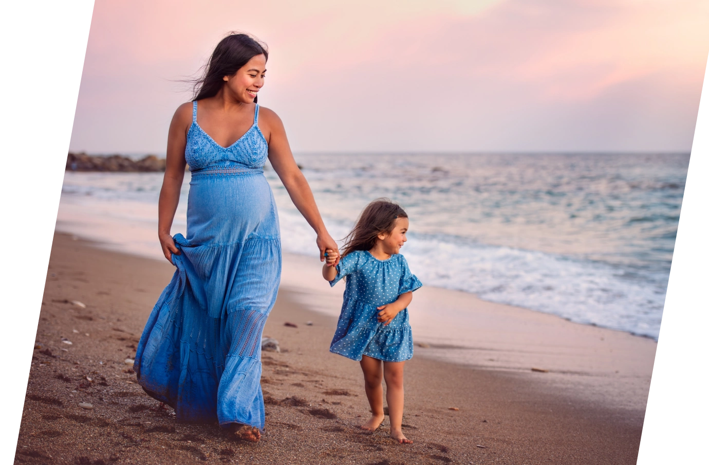 an expecting mom with travel insurance for pregnancy holds her daughter's hand as they walk down the beach at sunset