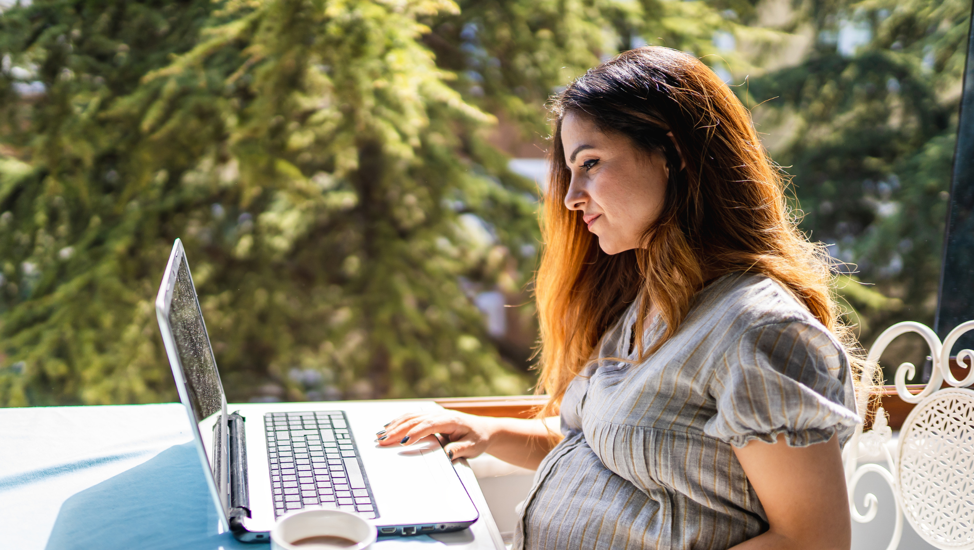 a pregnant woman sits on a laptop outdoors, looking up